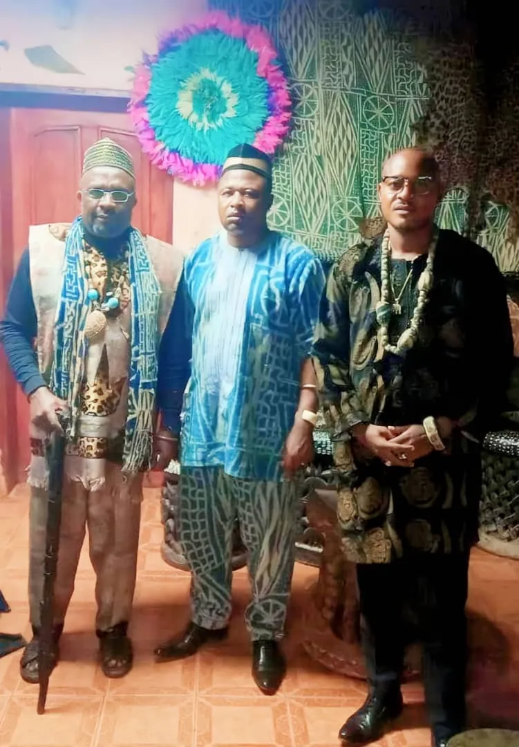 Trois hommes en tenues traditionnelles devant un mur orné de tissus ndop et d’un ornement de plumes, à la chefferie Bangoulap