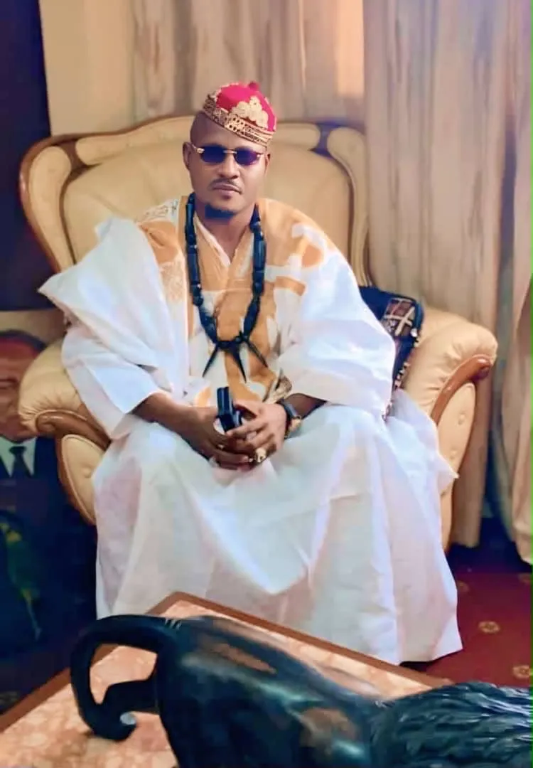 Chef en agbada blanc brodé or, bonnet rouge et collier de perles, assis dans un fauteuil