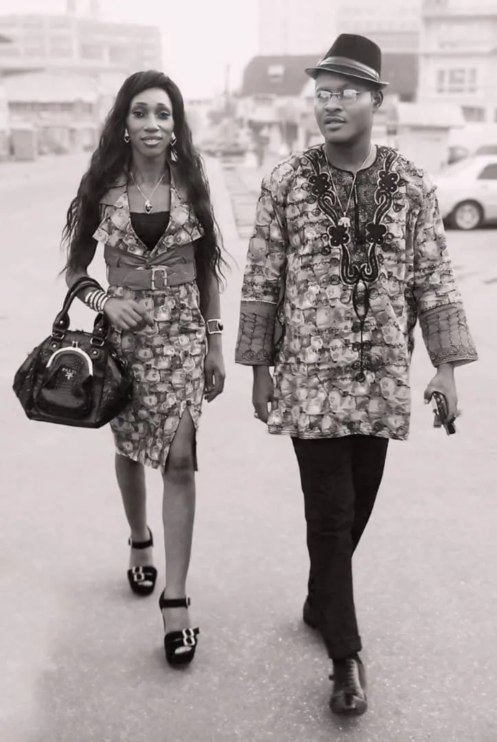 Portrait en noir et blanc de Joseph Kuika marchant en tenue brodée et chapeau, accompagné d’une invitée